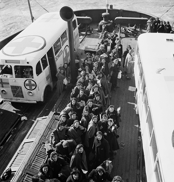 Group of people stand on a ship's deck looking up at the photographer. On the deck are also two white buses with Swedish flags and the red cross symbol.
