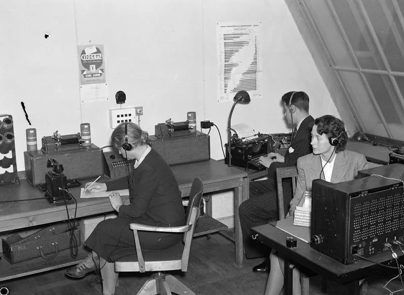 Tre personer sitter i ett rum med radioapparater. Alla tre har hörlurar på sig och verkar koncentrerade å sitt arbete 