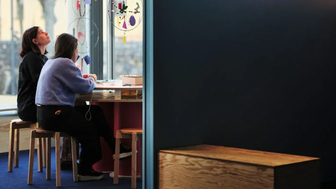 Två personer sitter på pallar vid ett bord i museet. Ovanför hänger installationer byggda med ståltråd och papper.