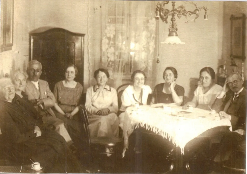 Foto från 1930-talet. En grupp människor sitter inomhus framför ett runt bord. De tittar in i kameran. Längst till vänster farmor Lina och längst till höger Veras föräldrar.