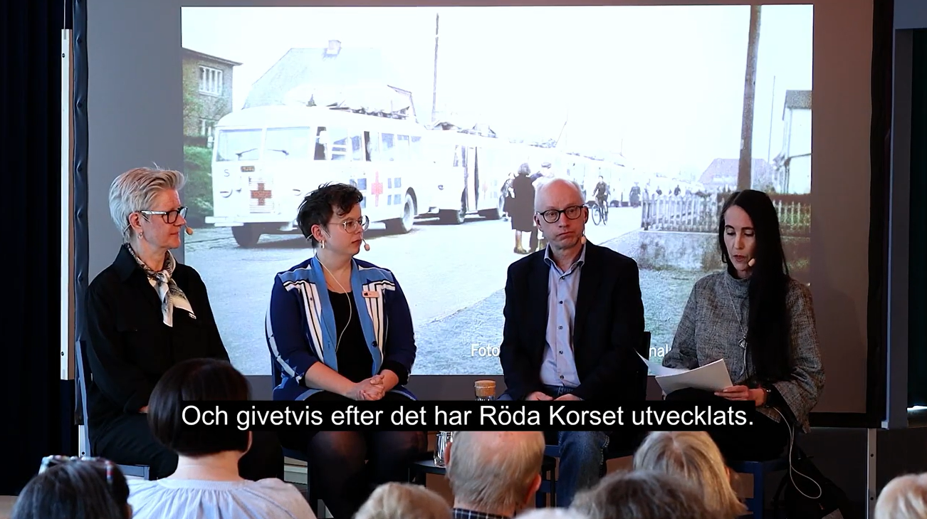 Fyra personer sittandes på en scen med en bild av vita bussar i bakgrunden